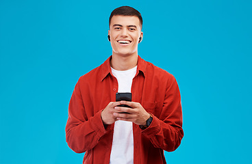 Image showing Man, earphones and smile with phone for communication in portrait, studio and happy by blue background. Asian male model, text and chat on smartphone while streaming music, song and audio or podcast