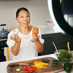 Image showing Asian woman, vegetables and live streaming for cooking with bottle, healthy food and blog on social media in kitchen. Chef, mature influencer and content creation, class and broadcast online tutorial