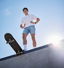 Image showing Blue sky, skateboard and young man from below on skate ramp ready for trick. Fitness, street fashion and urban sports, a skater in skatepark and summer sun for adventure, freedom and extreme sport.