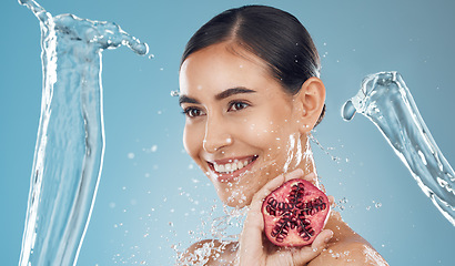 Image showing Woman, beauty and water splash with pomegranate, skincare and happy against a blue studio background. Model, smile and shower with fruit for fresh, clean and healthy skin, face or hair with happiness