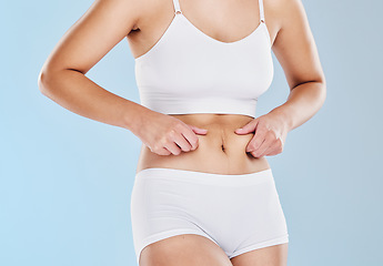 Image showing Plus size, fat and woman touching her stomach cellulite in underwear standing in a studio. Overweight, obesity and closeup of a girl model feeling her chubby abdomen isolated by a blue background.