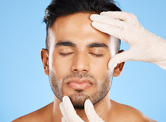 Image showing Cosmetic, plastic surgery and face of young man, for skincare and white gloves on blue studio background for dermatology care. Makeup, Indian male and skin rejuvenation for wellness and collagen.