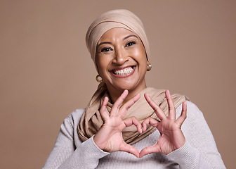 Image showing Islam woman, heart and sign with smile, happy and peace against brown studio background. Muslim lady, mature female and hand gesture for happiness, love and commitment for humanity and positive.