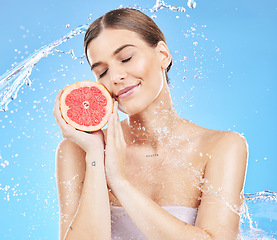 Image showing Woman, fruit and smile for skincare hydration, cosmetics or healthy nutrition against a blue studio background. Female smiling in satisfaction with water splash holding grapefruit for vitamin C skin