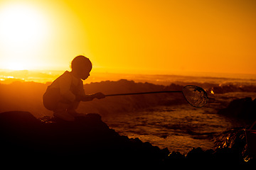 Image showing Sunset, silhouette and child on a rock at the beach playing or fishing with net in the water. Travel, seaside and girl kid in nature by the ocean in the evening to play or have fun while on vacation.