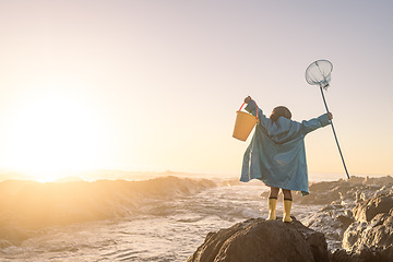 Image showing Child, freedom and fishing at sunset while at beach excited, happy and waiting for ocean waves outdoor while on vacation. Girl on a rock with a bucket and net to catch fish and have fun on holiday