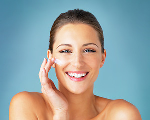 Image showing Dermatology, portrait and happy woman with cream on face, cosmetics and product for makeup on blue background. Happiness, skincare and wellness for model with smile, beauty and lotion for facial glow