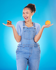 Image showing Portrait, excited and woman with donut, orange and choice in studio isolated on blue background. Happy, fruit or person with doughnut, fast food or comparison for healthy diet, nutrition or wellness