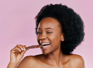 Image showing Black woman, eating chocolate and diet with candy and beauty, health and dessert snack on pink background. Skin, glow and female model wink with cacao sweets, calories and cosmetics in studio