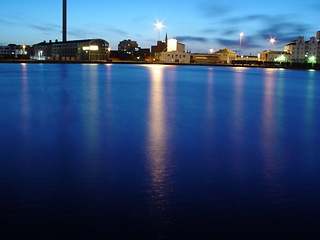 Image showing Ocean, night and buildings in port with lights, reflection on calm blue water or holiday location. City at sea in evening, travel, skyline and nature, urban beach village for vacation, peace and view