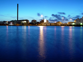 Image showing Sea, night and buildings in port with lights, reflection on calm blue water or holiday location. City at ocean in evening, travel, skyline and nature, urban beach village for vacation, peace and view