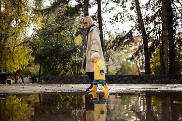 Image showing Small bond infant boy wearing yellow rubber boots and yellow waterproof raincoat walking in puddles on a overcast rainy day holding her mother's hand. Mom with small child in rain outdoors.
