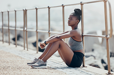 Image showing Beach, relax and woman runner thinking after an outdoor cardio workout in nature by ocean. Tired African athlete, running and fitness girl sitting on promenade to breathe, calm and rest after workout