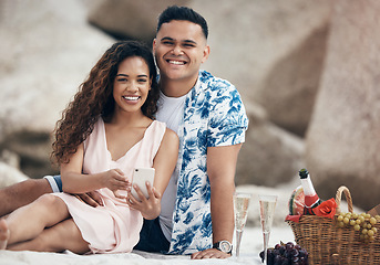 Image showing Couple, phone and beach picnic date with champagne, food basket and fruit for anniversary celebration, valentines day or love bonding. Portrait, smile or happy woman with relax man and 5g mobile tech