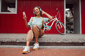 Image showing Happy woman, phone and headphones for music or listening to podcast while sitting on a urban city street singing a song with a bike to travel. Female using technology to stream audio on a sidewalk