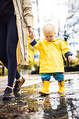 Image showing Small bond infant boy wearing yellow rubber boots and yellow waterproof raincoat walking in puddles on a overcast rainy day holding her mother's hand. Mom with small child in rain outdoors.