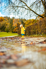 Image showing Sun always shines after the rain. Small bond infant boy wearing yellow rubber boots and yellow waterproof raincoat walking in puddles in city park on sunny rainy day.