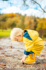 Image showing Sun always shines after the rain. Small bond infant boy wearing yellow rubber boots and yellow waterproof raincoat walking in puddles in city park on sunny rainy day.