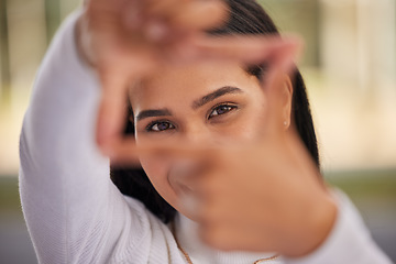 Image showing Hands, frame and eyes with a woman looking through her fingers while framing her face outdoor. Portrait, girl and happy with a young female posing behind a hand sign on a blurry background outside