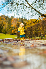 Image showing Sun always shines after the rain. Small bond infant boy wearing yellow rubber boots and yellow waterproof raincoat walking in puddles in city park on sunny rainy day.