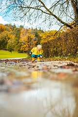 Image showing Sun always shines after the rain. Small bond infant boy wearing yellow rubber boots and yellow waterproof raincoat walking in puddles in city park on sunny rainy day.