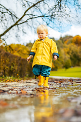 Image showing Sun always shines after the rain. Small bond infant boy wearing yellow rubber boots and yellow waterproof raincoat walking in puddles in city park on sunny rainy day.