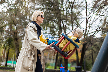 Image showing Mother pushing her infant baby boy child wearing yellow rain boots and cape on swing on playground outdoors on cold rainy overcast autumn day in Ljubljana, Slovenia