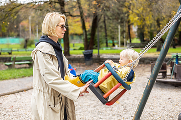 Image showing Mother pushing her infant baby boy child wearing yellow rain boots and cape on swing on playground outdoors on cold rainy overcast autumn day in Ljubljana, Slovenia