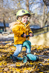 Image showing Adorable toddler boy wearing yellow protective helmet riding baby scooter outdoors on autumn day. Kid training balance on mini bike in city park. Fun autumn outdoor activity for small kids.