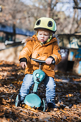 Image showing Adorable toddler boy wearing yellow protective helmet riding baby scooter outdoors on autumn day. Kid training balance on mini bike in city park. Fun autumn outdoor activity for small kids.