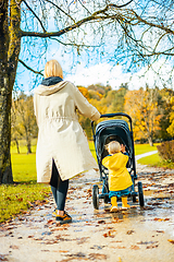 Image showing Sun always shines after the rain. Small blond infant boy wearing yellow rubber boots and yellow waterproof raincoat walking in puddles, pushing stroller in city park, holding mother's hand after rain.