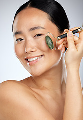 Image showing Asian woman, cosmetics and facial roller for wellness, skincare and smooth face with studio background. Young female, girl and anti aging for natural beauty, smile and happy for health and body care.