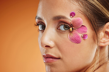 Image showing Model, flower and eye for makeup at studio for art, portrait and natural beauty. Woman, flowers and eyes with floral organic cosmetics on skin with glow, shine and healthy face with orange background