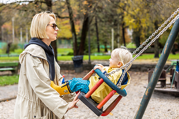 Image showing Mother pushing her infant baby boy child wearing yellow rain boots and cape on swing on playground outdoors on cold rainy overcast autumn day in Ljubljana, Slovenia