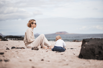 Image showing Mother enjoying winter beach vacations playing with his infant baby boy son on wild volcanic sandy beach on Lanzarote island, Canary Islands, Spain. Family travel and vacations concept