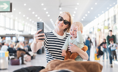 Image showing Mother taking selfie with mobile phone, while traveling with child, holding his infant baby boy at airport, waiting to board a plane. Travel with kids concept.