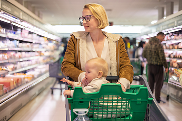 Image showing Mother pushing shopping cart with her infant baby boy child down department aisle in supermarket grocery store. Shopping with kids concept.