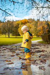 Image showing Sun always shines after the rain. Small bond infant boy wearing yellow rubber boots and yellow waterproof raincoat walking in puddles in city park on sunny rainy day.