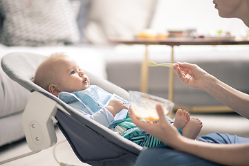 Image showing Mother spoon feeding her baby boy infant child in baby chair with fruit puree. Baby solid food introduction concept.