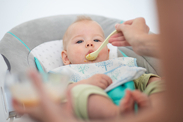 Image showing Mother spoon feeding her baby boy infant child in baby chair with fruit puree. Baby solid food introduction concept.