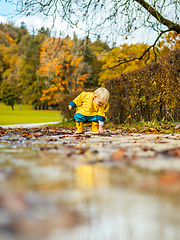 Image showing Sun always shines after the rain. Small bond infant boy wearing yellow rubber boots and yellow waterproof raincoat walking in puddles in city park on sunny rainy day.