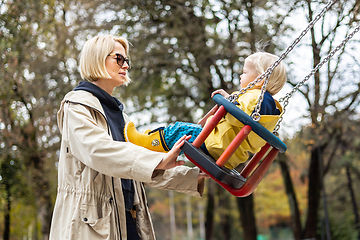 Image showing Mother pushing her infant baby boy child wearing yellow rain boots and cape on swing on playground outdoors on cold rainy overcast autumn day in Ljubljana, Slovenia