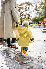 Image showing Small bond infant boy wearing yellow rubber boots and yellow waterproof raincoat walking in puddles on a overcast rainy day holding her mother's hand. Mom with small child in rain outdoors.