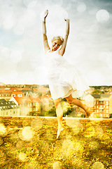 Image showing Portrait, freedom and jump with a woman on a roof in the city and fun against a bokeh effect. Smile, neighborhood or arms raised and a happy young person with energy outdoor to dance in summer