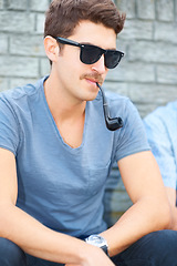 Image showing Man in sunglasses, smoking pipe and thinking, relax outdoor and fashion in urban city by wall. Smoke, cool person in glasses or style, serious or hipster in casual clothes, tshirt and sitting in town