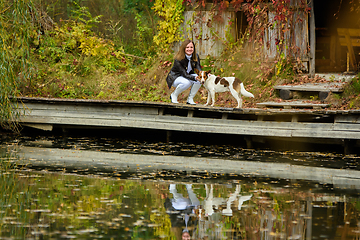 Image showing A young beautiful girl walks with a dog near a lake in an autumn park
