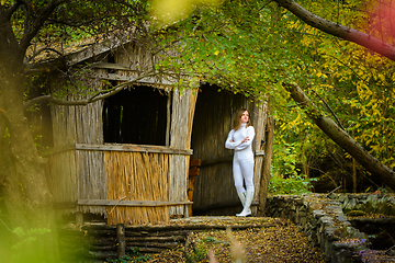 Image showing A young beautiful girl in white clothes stands near an old abandoned house in the forest