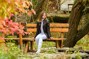 Image showing A young beautiful girl sits on a bench in an autumn park and looks at the surrounding nature