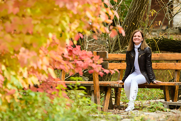 Image showing A young beautiful girl sits on a bench in an autumn park and looks happily into the frame