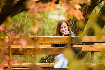 Image showing A young beautiful girl sits on a bench in an autumn park and looks into the frame, smiling.